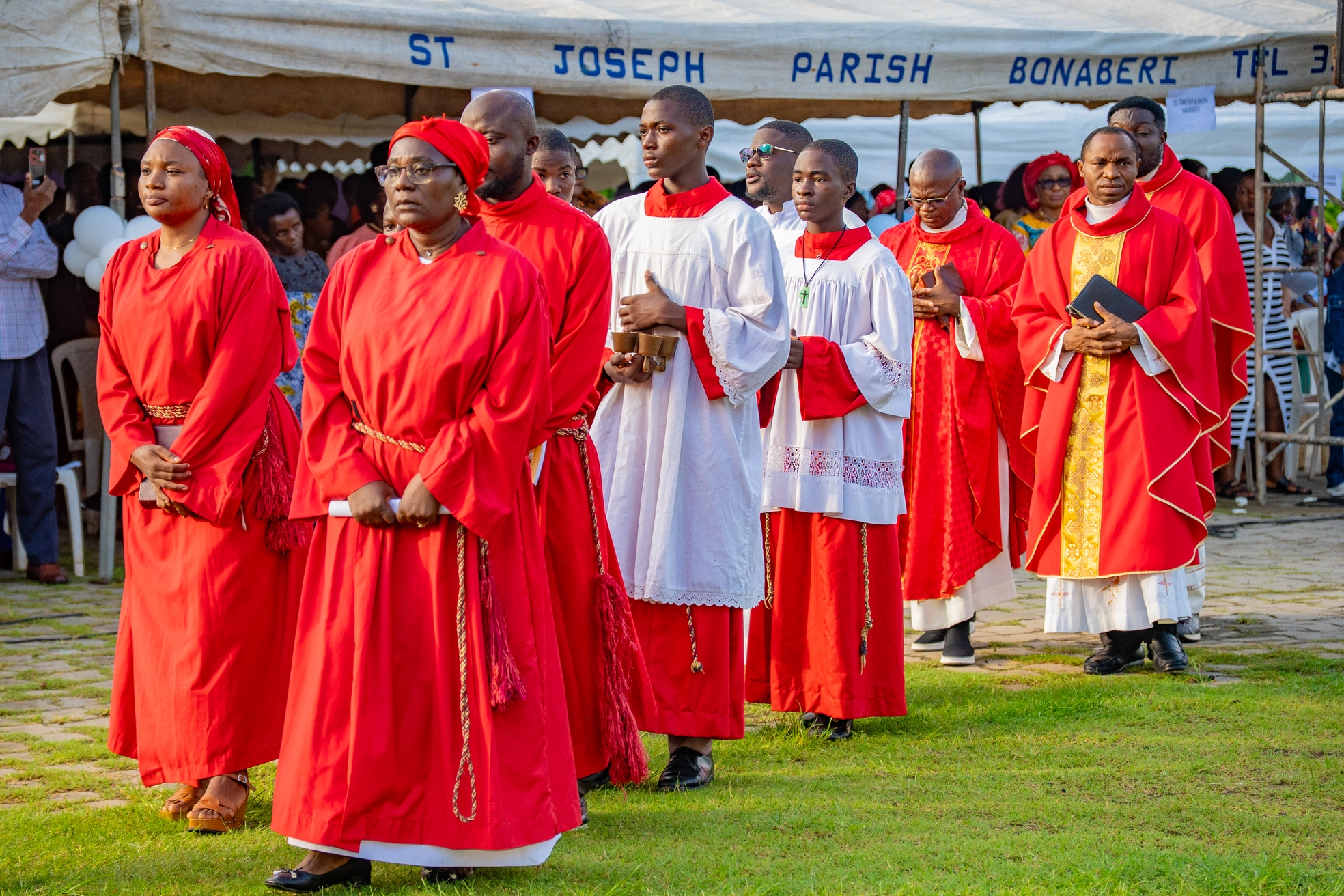Clergy and choir in red vestments processing past the parish canopy